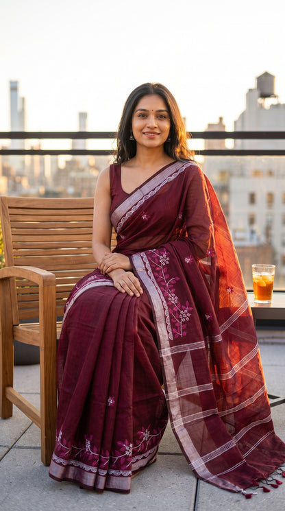 Model seated comfortably on a wooden chair on a rooftop terrace during sunset, showcasing the soft texture, neat pleats, and silver Zari highlights of a maroon cotton linen saree