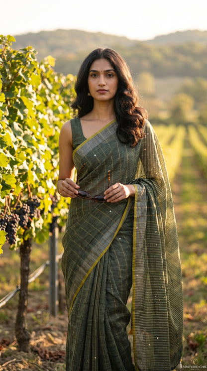 A model holding sunglasses while standing in a sunlit afternoon vineyard, showcasing the straight silhouette, subtle micro-sequin sparkle, and contrast yellow trim of an olive green khadi cotton linen saree