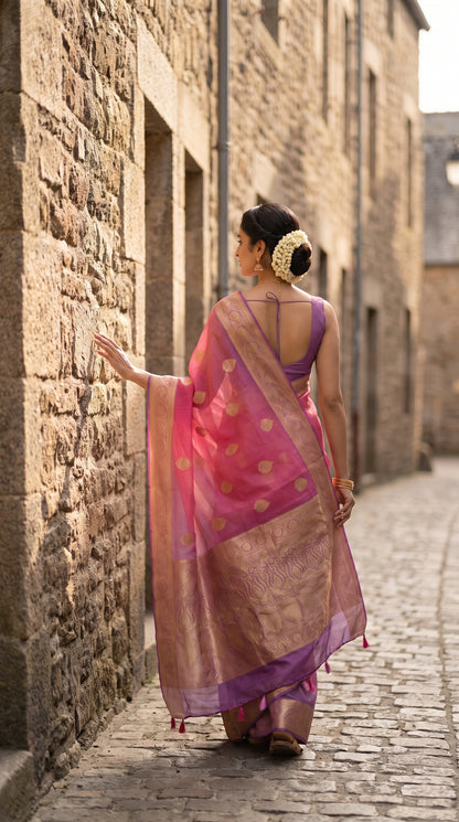 A rear-view portrait of a woman walking along a historic cobblestone lane at Mont Saint-Michel, displaying the flowing purple zari pallu with hand-tied tassels trailing behind her pink pure Kora silk saree