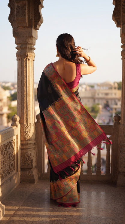 A rear-view portrait of a woman with her hair in a bun looking out from a sandstone balcony palace terrace, displaying the sprawling, heavily woven multicolored jamawar pallu of her black silk saree