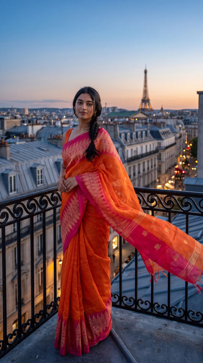 A full-length portrait of a woman standing on a classic black wrought-iron balcony overlooking Paris rooftops at sunset, showcasing her flowing orange linen saree pallu catching the breeze with the Eiffel Tower in the background