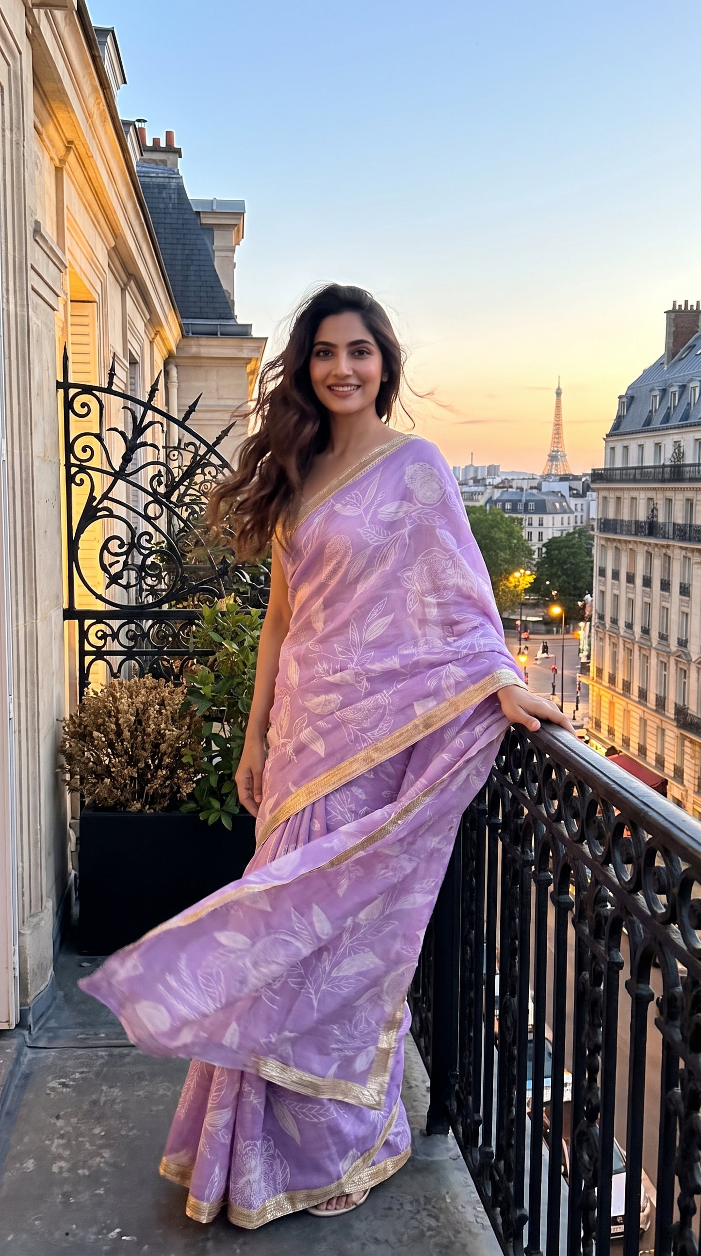 A portrait of a smiling woman standing on a Paris balcony at twilight with the Eiffel Tower in the background, showing the breezy movement and translucent texture of her lavender printed muslin saree