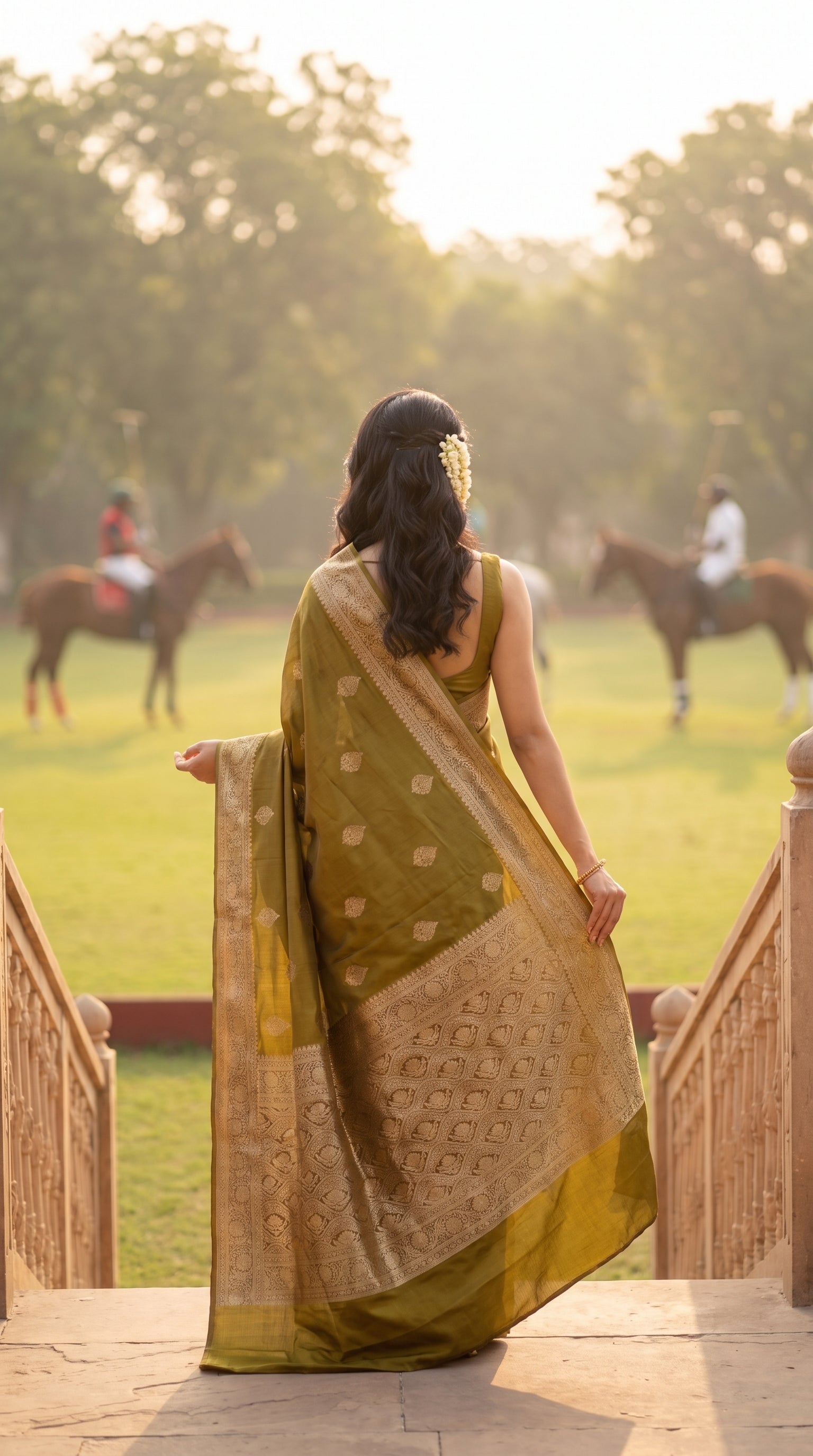 A rear view of a woman with a gajra in her hair descending a palace terrace staircase, looking out towards a polo field with horses, displaying the heavily woven gold zari work across the pallu of her olive green saree