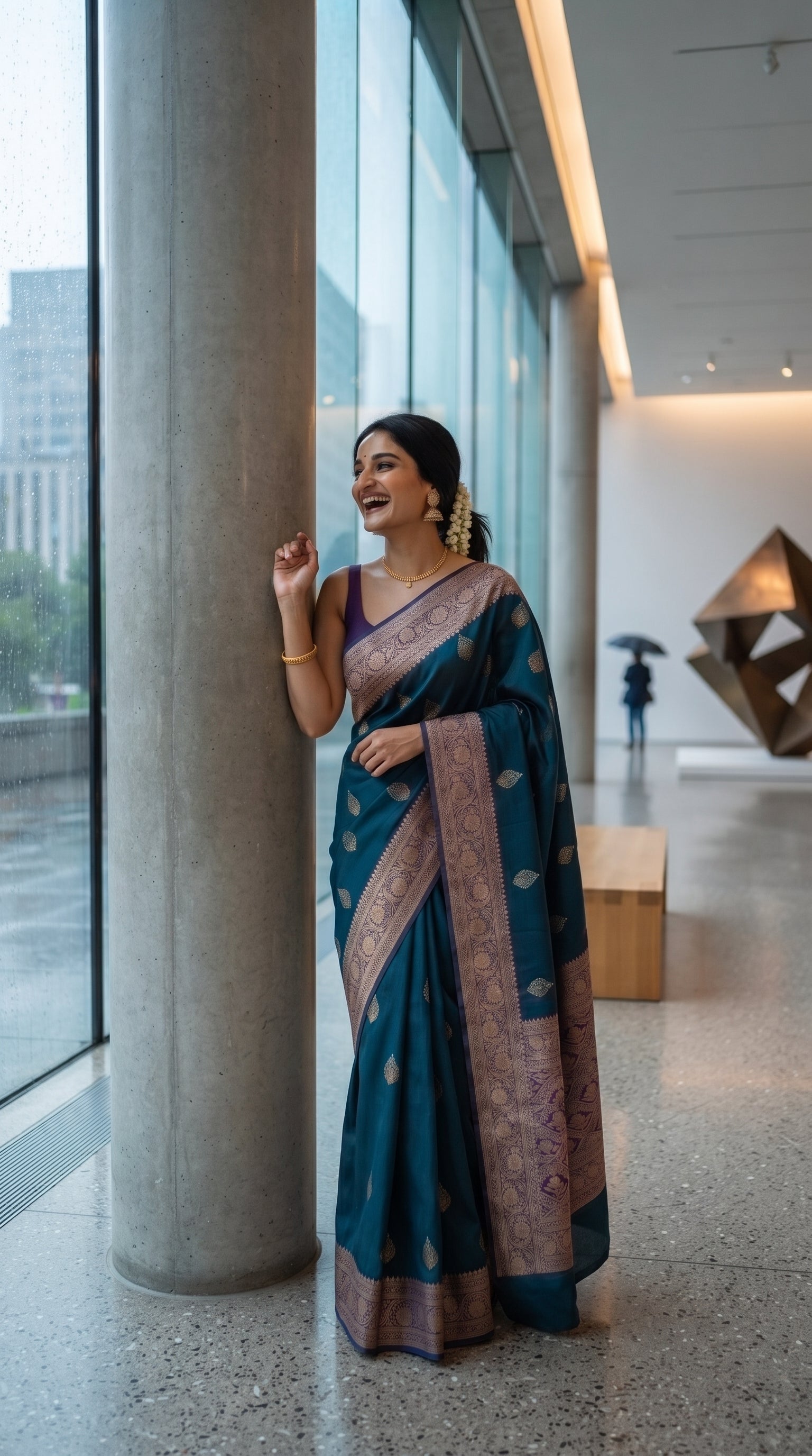 A full-length portrait of a laughing woman standing next to a concrete pillar inside a modern glass museum building, showcasing the rich drape and gold-woven borders of her teal silk saree