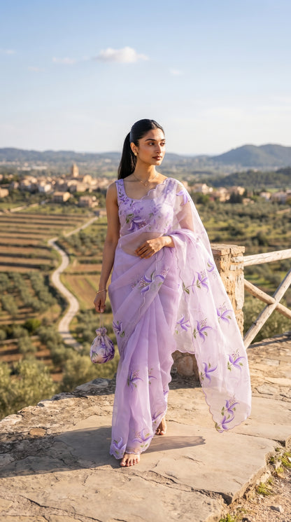 A full-length portrait of a woman standing at a stone wall viewpoint overlooking a scenic countryside road, beautifully displaying the fluid movement, sheer texture, and scalloped cutwork borders of her lavender chiffon saree while holding a matching potli bag