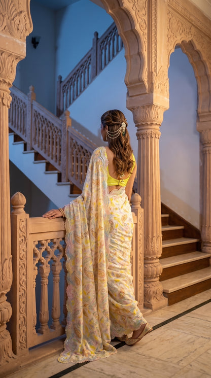 A rear-view portrait of a woman looking down a grand indoor stone staircase from a balcony railing, displaying her detailed hair decoration and the cascading cutwork border of her cream silk saree
