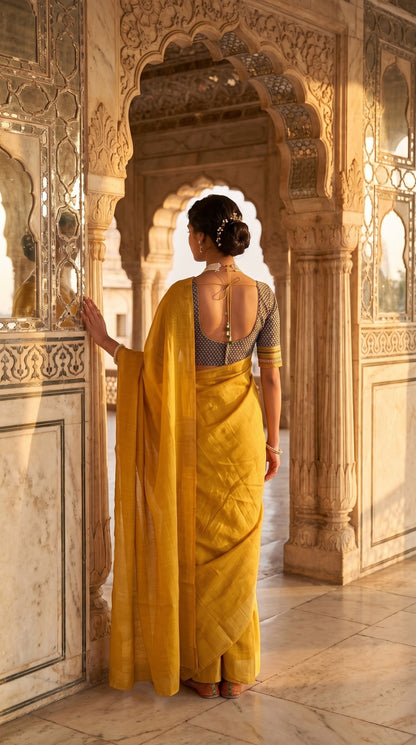 A rear portrait of a woman standing in a majestic palace archway, showing the tie-back detailing of her dark geometric print blouse and the graceful translucent cascade of her yellow-golden tissue cotton saree