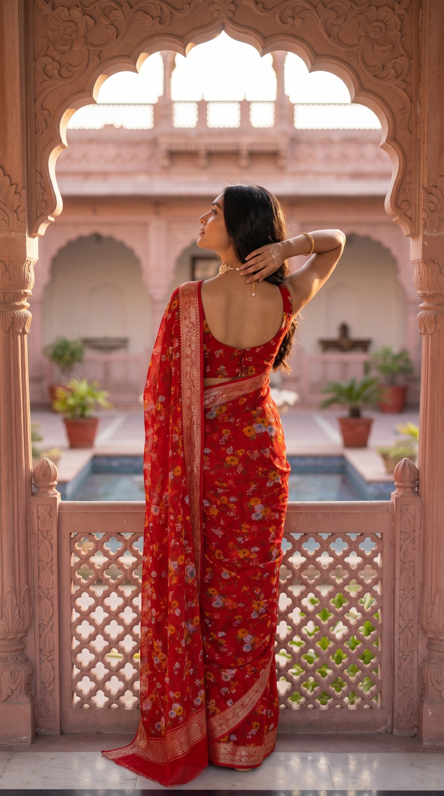 An over-the-shoulder back view of a woman standing by an ornate stone palace window balcony, highlighting the sleek backline of her sleeveless red blouse and the detailed jacquard border pattern