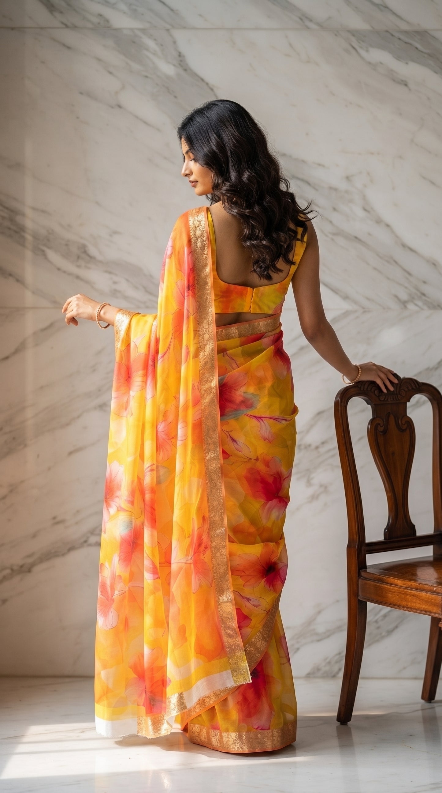 A rear-view portrait of a woman with long wavy hair looking back over her shoulder while resting her hand on a wooden chair, showcasing the sleeveless blouse design and translucent pallu of her yellow and orange floral printed saree