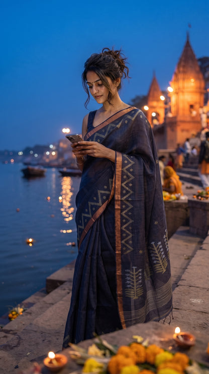 A young woman checks her phone while standing on a river ghat at twilight with glowing temples behind her, elegantly displaying the copper-gold zigzag Zari borders on her charcoal blue tissue linen saree