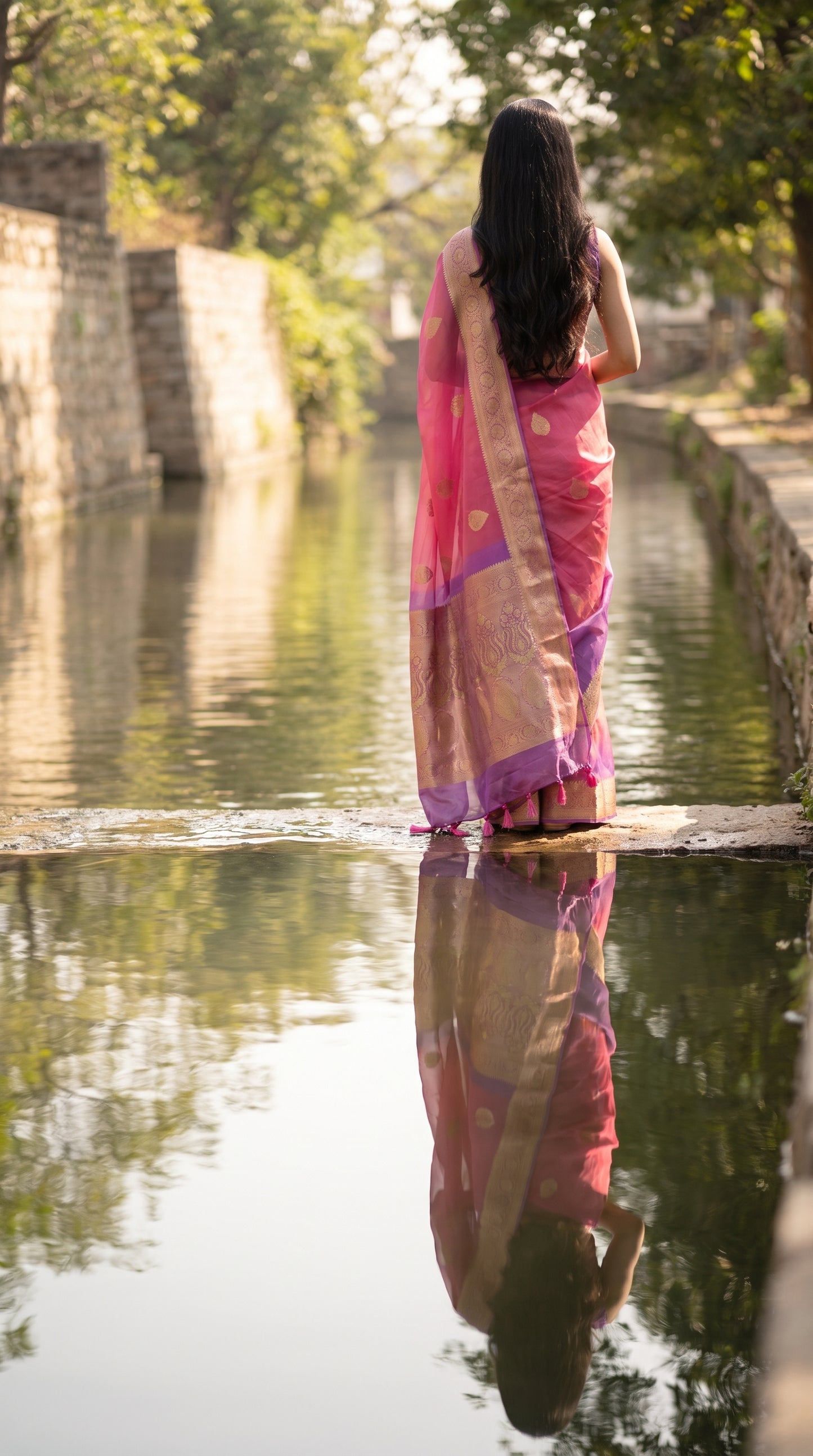 A rear portrait of a woman standing by a quiet stone canal path, with her translucent purple and pink silk saree pallu reflecting beautifully in the clear water surface