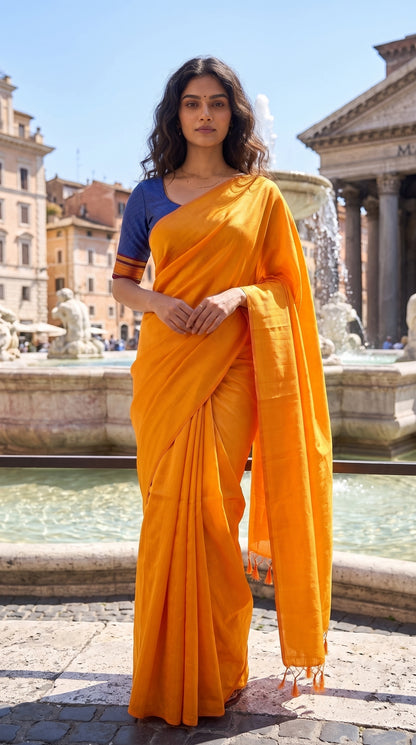 A full-length front portrait of a woman standing near a grand historic stone fountain, showcasing the clean structured pleats, solid canvas, and subtle golden-orange glaze of her tissue cotton saree paired with a blue blouse
