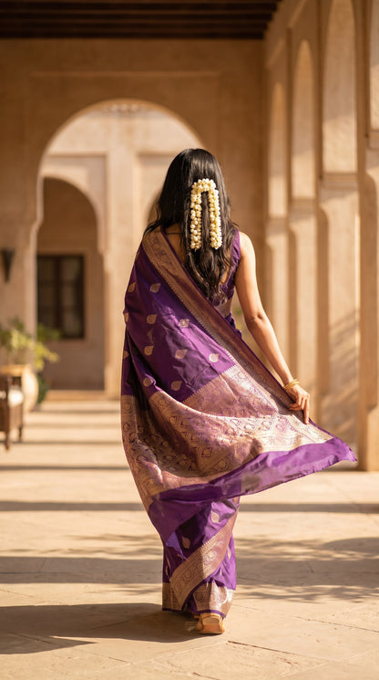 A rear-view portrait of a woman with a gajra hair decoration walking down a sun-drenched sandstone arched corridor, showcasing the flowing drape and gold-woven pallu patterns of her royal purple silk saree