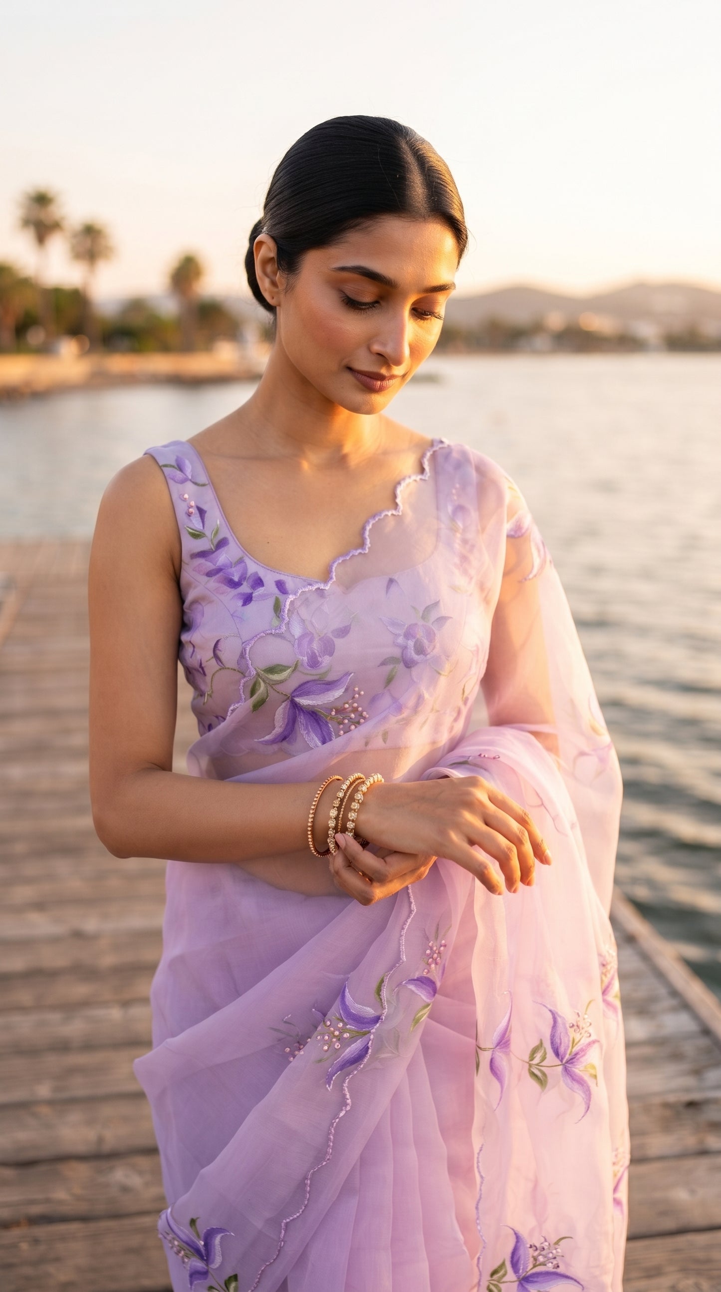 A close-up shot of a woman with a sleek low bun standing on a wooden jetty at sunrise, adjusting her gold bangles, showcasing the delicate cutwork neckline and floral detailing on her lavender blouse