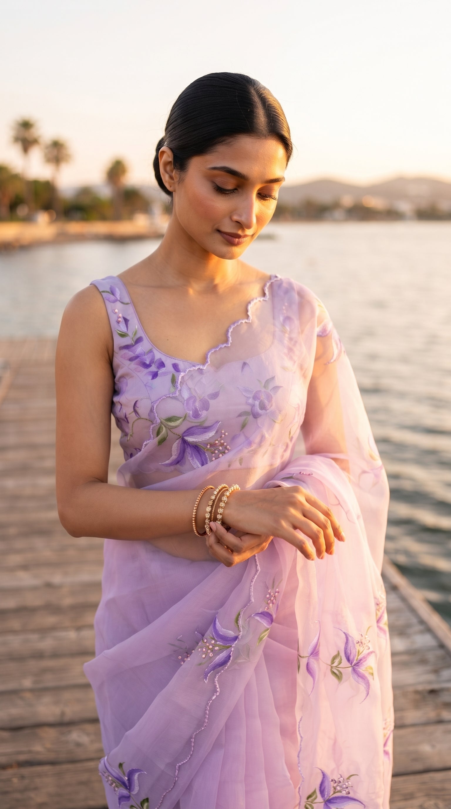 A close-up shot of a woman with a sleek low bun standing on a wooden jetty at sunrise, adjusting her gold bangles, showcasing the delicate cutwork neckline and floral detailing on her lavender blouse