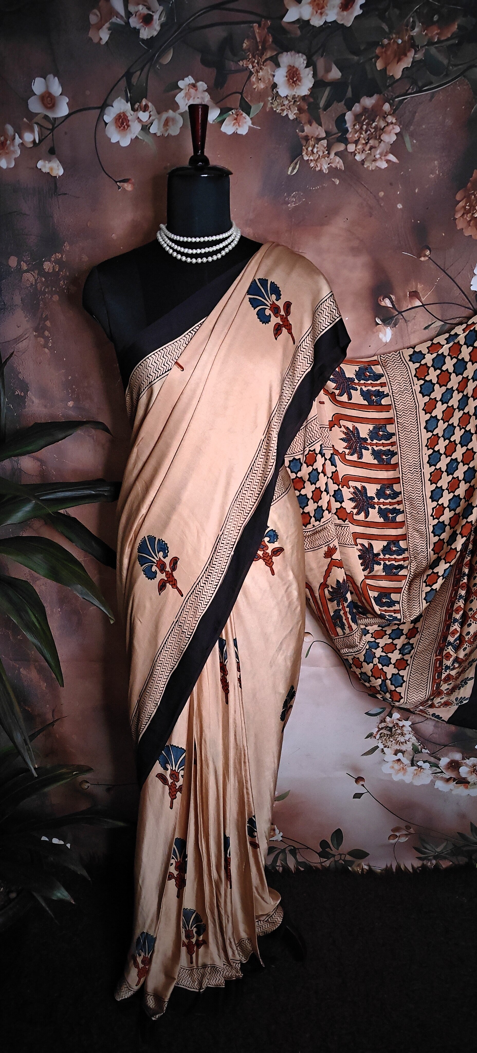 Ajrakh Modal Silk Saree – Beige with Black Borders and Blue-Maroon Pallu saree with floral patterns on a mannequin against a floral background