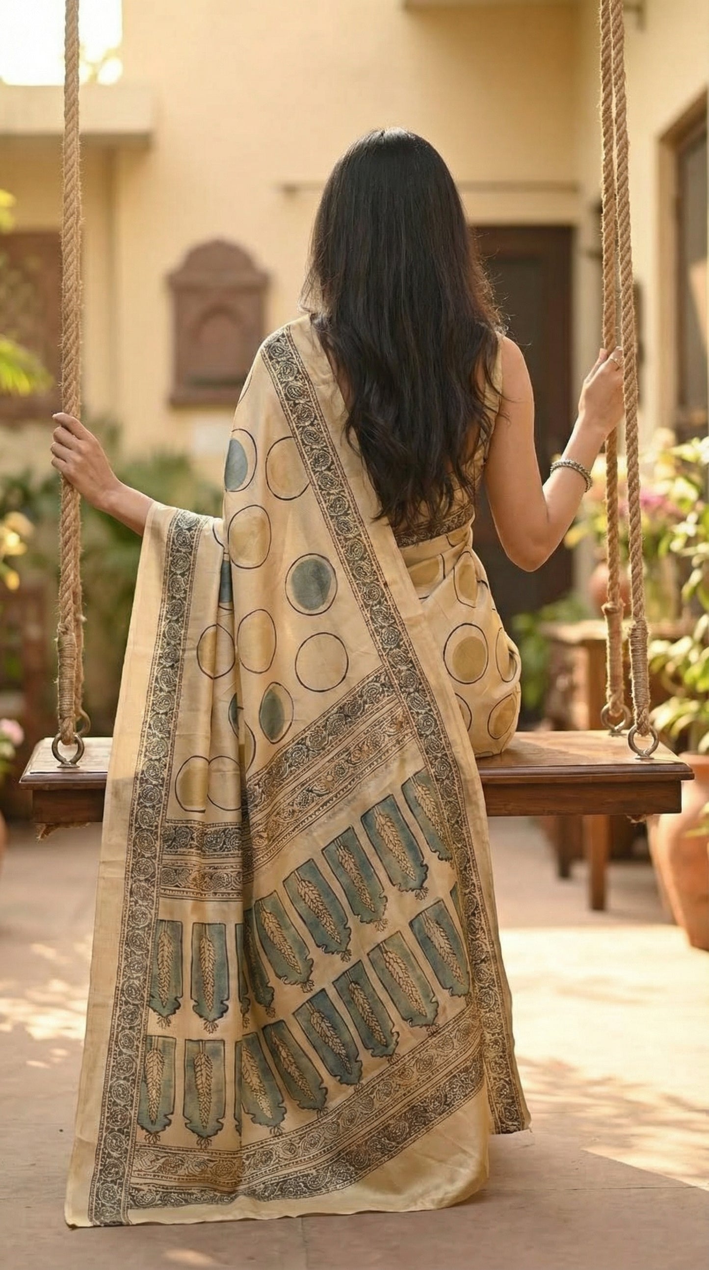 Woman wearing Ajrakh Modal Silk Saree – Beige with Golden & Olive Circular Motifs and Feather Pallu with intricate patterns sitting on a swing outdoors.