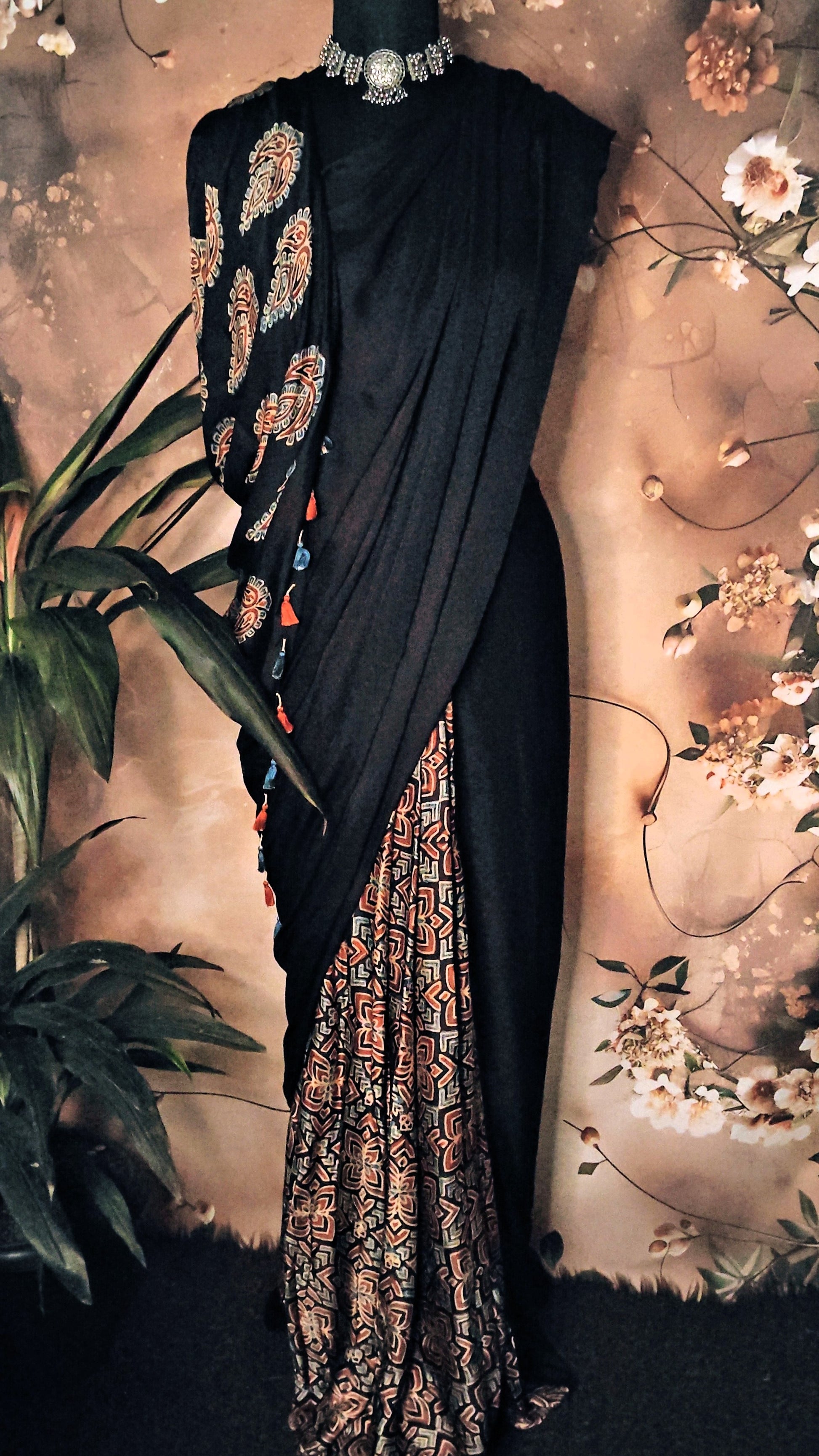 Ajrakh Modal Silk Saree – Black with Earthy Motifs and Tassel-Embellished Pallu saree with floral patterns on a decorative background.