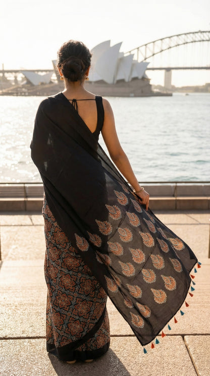 Woman wearing a Ajrakh Modal Silk Saree – Black with Earthy Motifs and Tassel-Embellished Pallu saree with floral patterns, with Sydney Opera House in the background.
