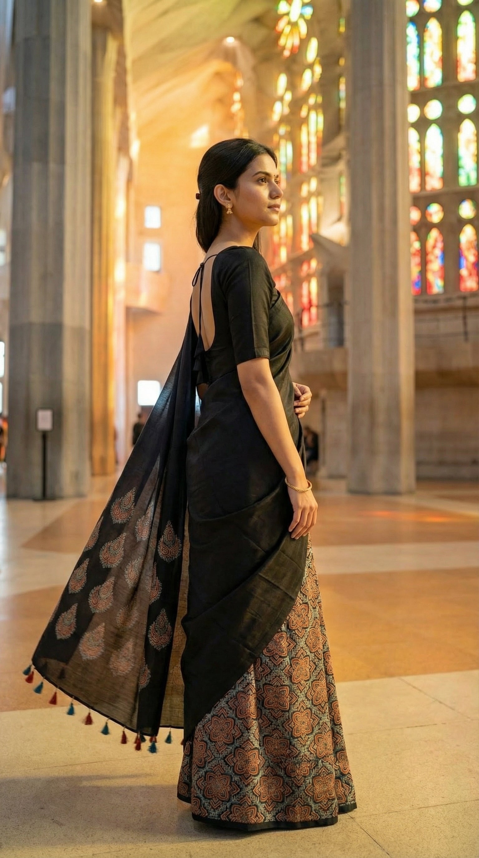 Woman wearing a Ajrakh Modal Silk Saree – Black with Earthy Motifs and Tassel-Embellished Pallu saree with floral patterns, standing in a cathedral with stained glass windows.