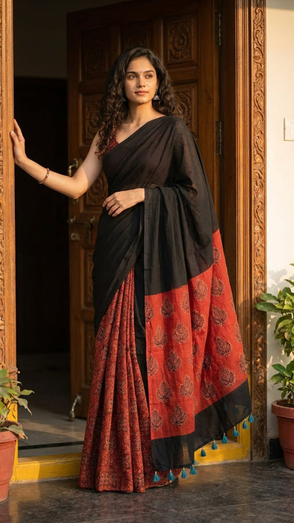 Woman wearing a ajrakh modal silk saree – black with red tribal motif pallu and geometric pleats saree draped standing in a doorway.