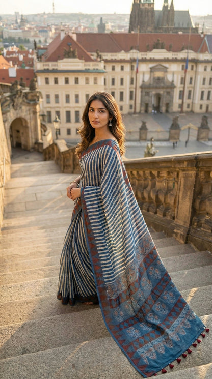 Woman in a Ajrakh Modal Silk Saree – Blue & Maroon with White Floral Painted and Striped Pallu saree with floral and striped patterns, standing on stone steps with a cityscape in the background.