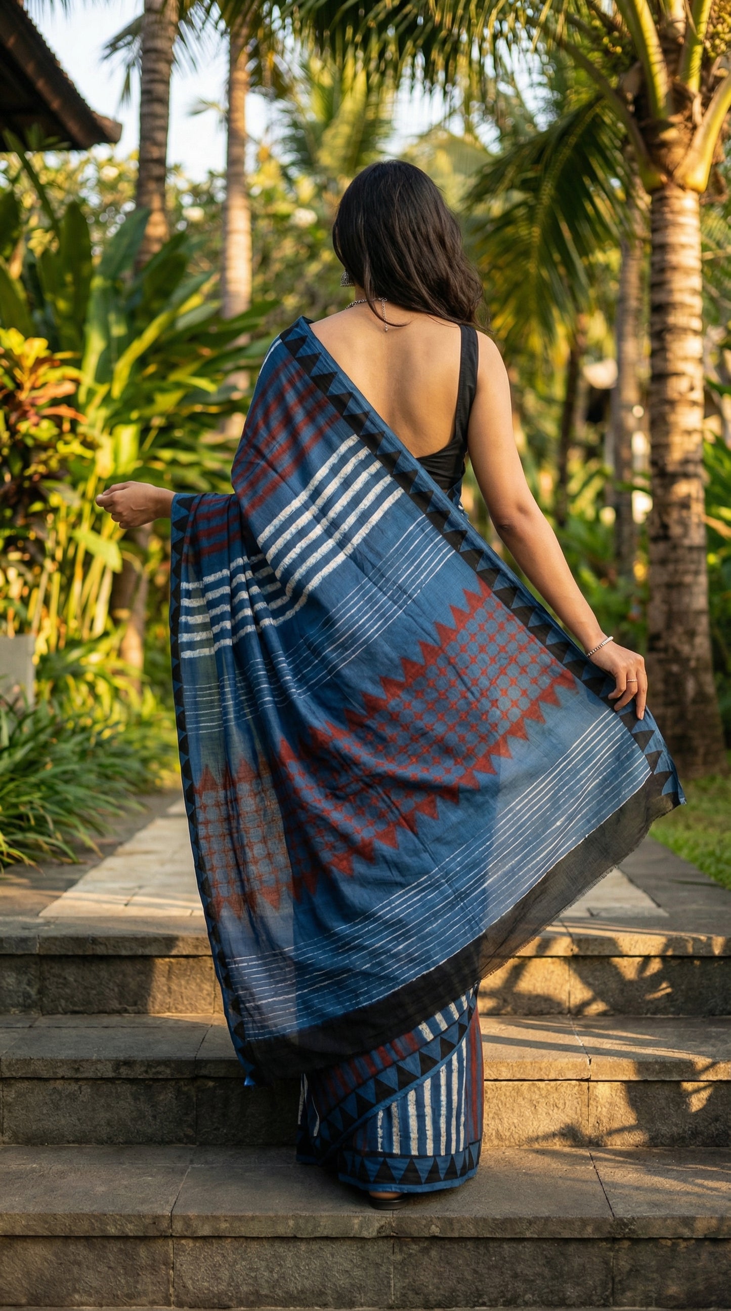Woman wearing a Ajrakh Modal Silk Saree – Deep Blue with Maroon Chevron & Triangular Border Motifs with a pattern standing on steps with palm trees in the background.