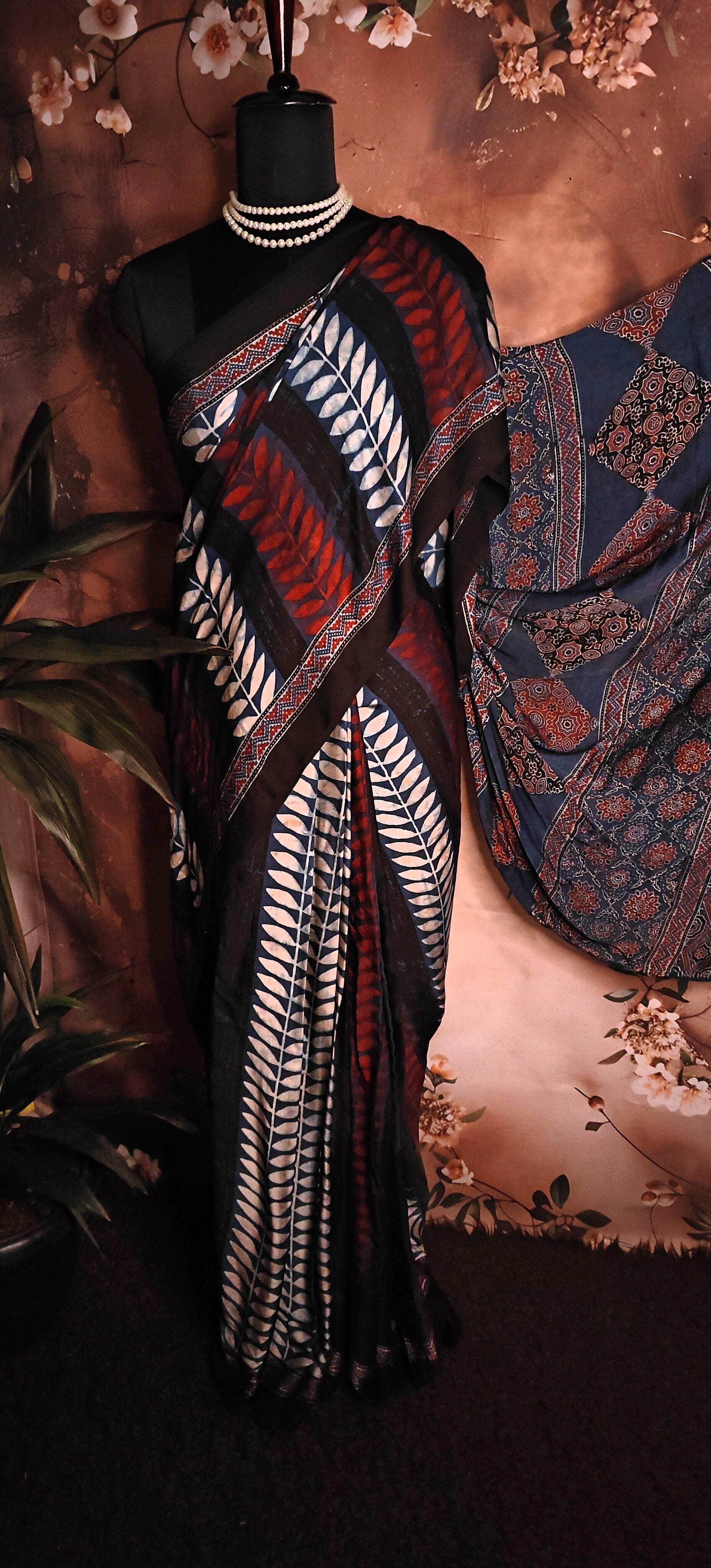 Ajrakh Modal Silk Saree – Geometric Black, White, Red & Blue with Intricate Pallu saree draped over a mannequin with a floral background