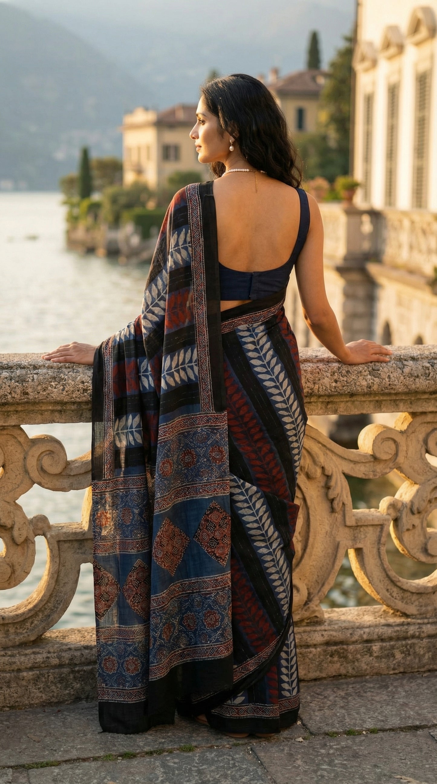Woman wearing a Ajrakh Modal Silk Saree – Geometric Black, White, Red & Blue with Intricate Pallu, standing on a balcony overlooking a scenic view with mountains and water.