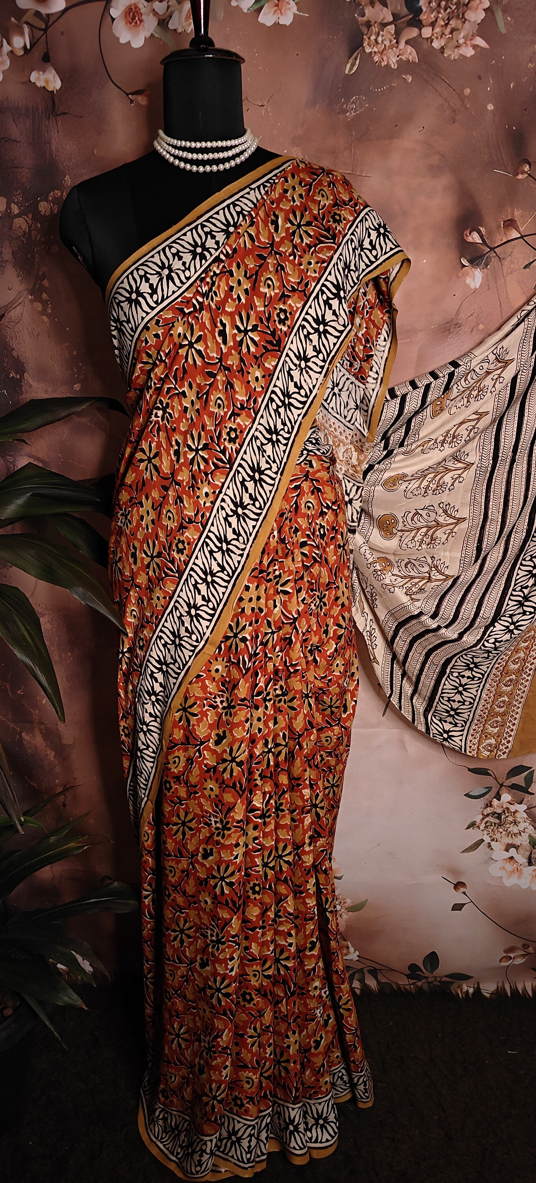Ajrakh Modal Silk Saree – Reddish-Orange with Black & White Border and Golden Pallu Motifs saree with intricate patterns on a mannequin against a floral background