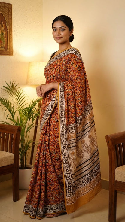 Woman wearing a Ajrakh Modal Silk Saree – Reddish-Orange with Black & White Border and Golden Pallu Motifs with intricate patterns in a home setting with a lamp and plant in the background.