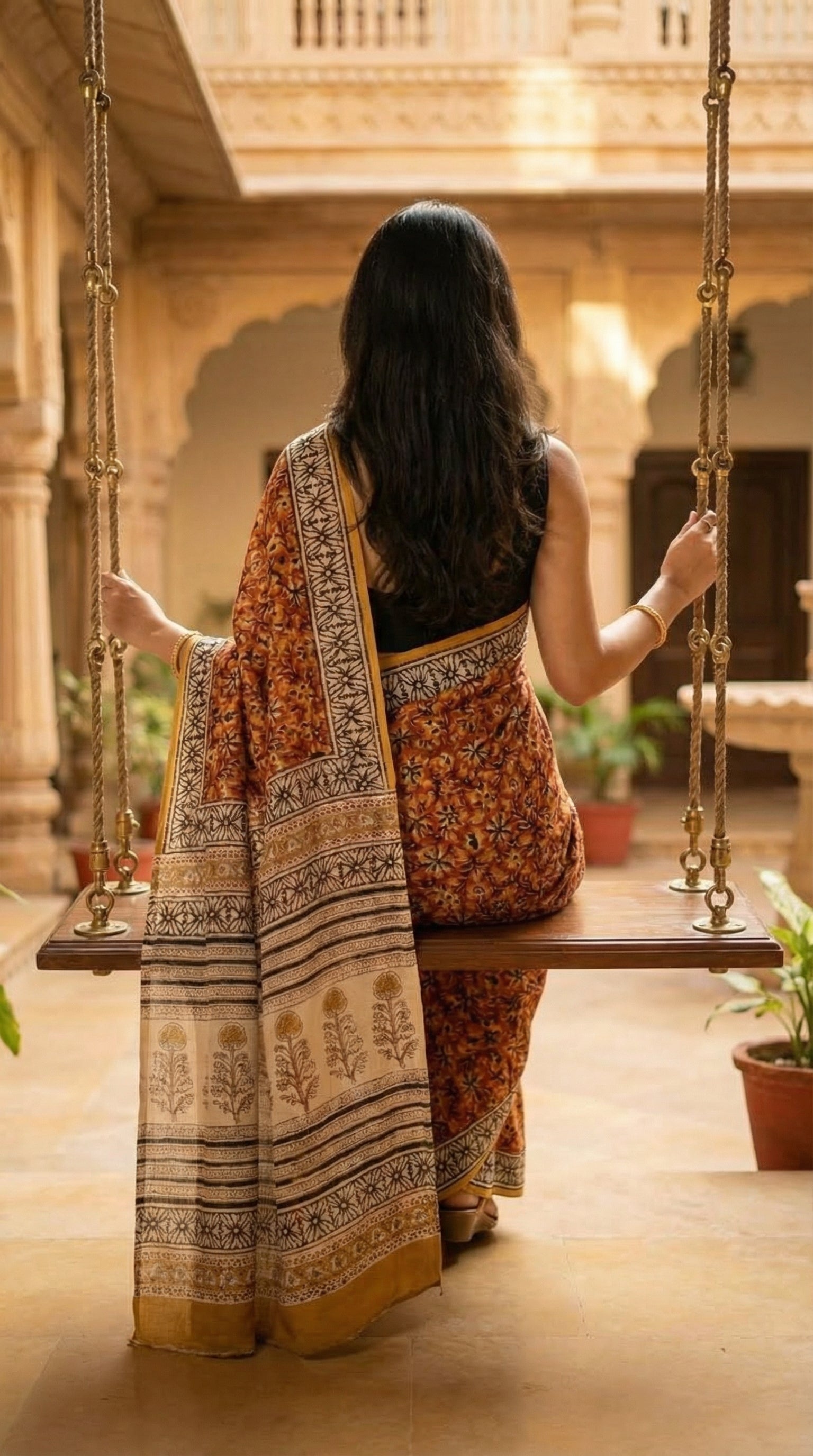 Woman wearing a Ajrakh Modal Silk Saree – Reddish-Orange with Black & White Border and Golden Pallu Motifs with intricate patterns sitting on a swing in a warm, sunlit indoor setting.