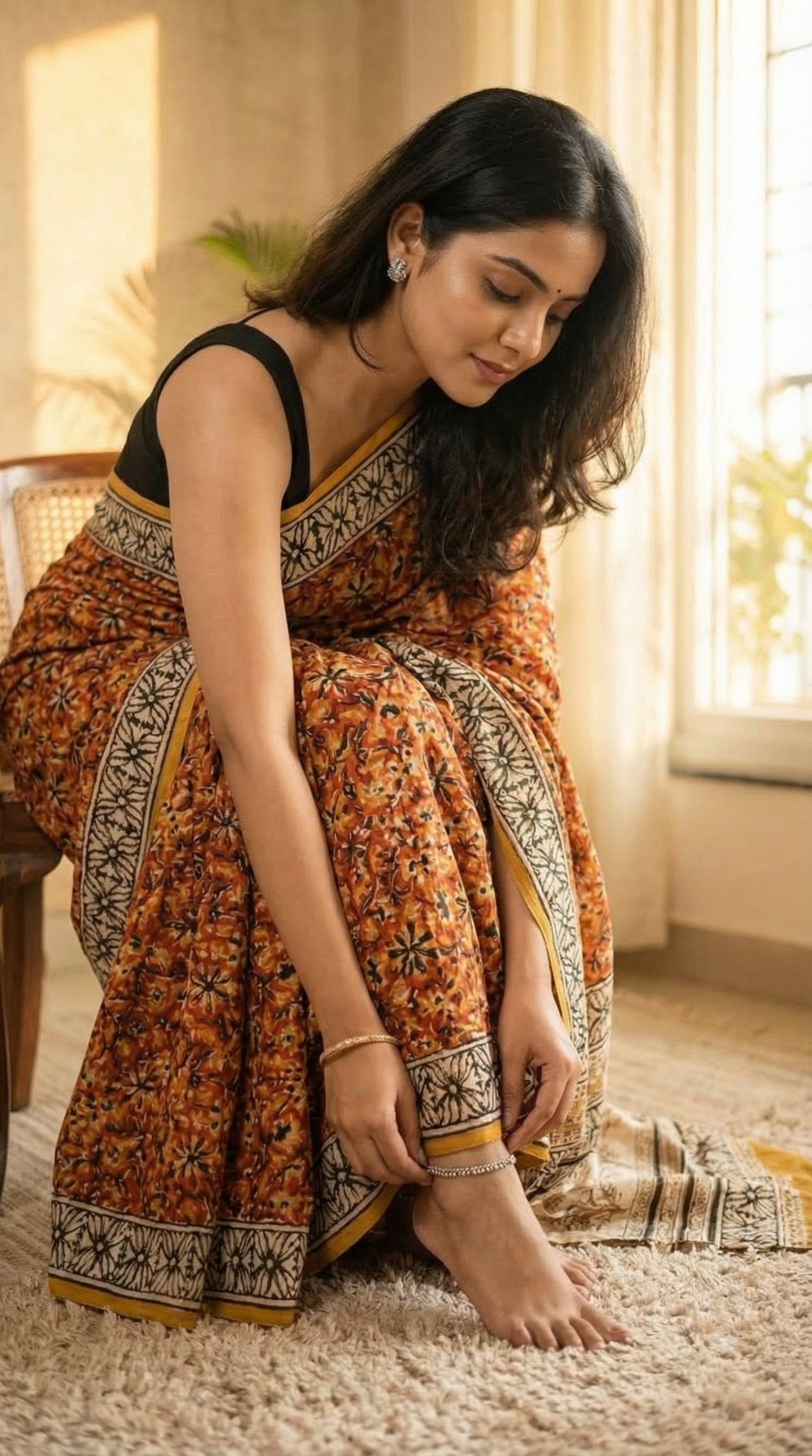 Woman wearing a Ajrakh Modal Silk Saree – Reddish-Orange with Black & White Border and Golden Pallu Motifs with intricate patterns sitting on a couch in a well-lit room.