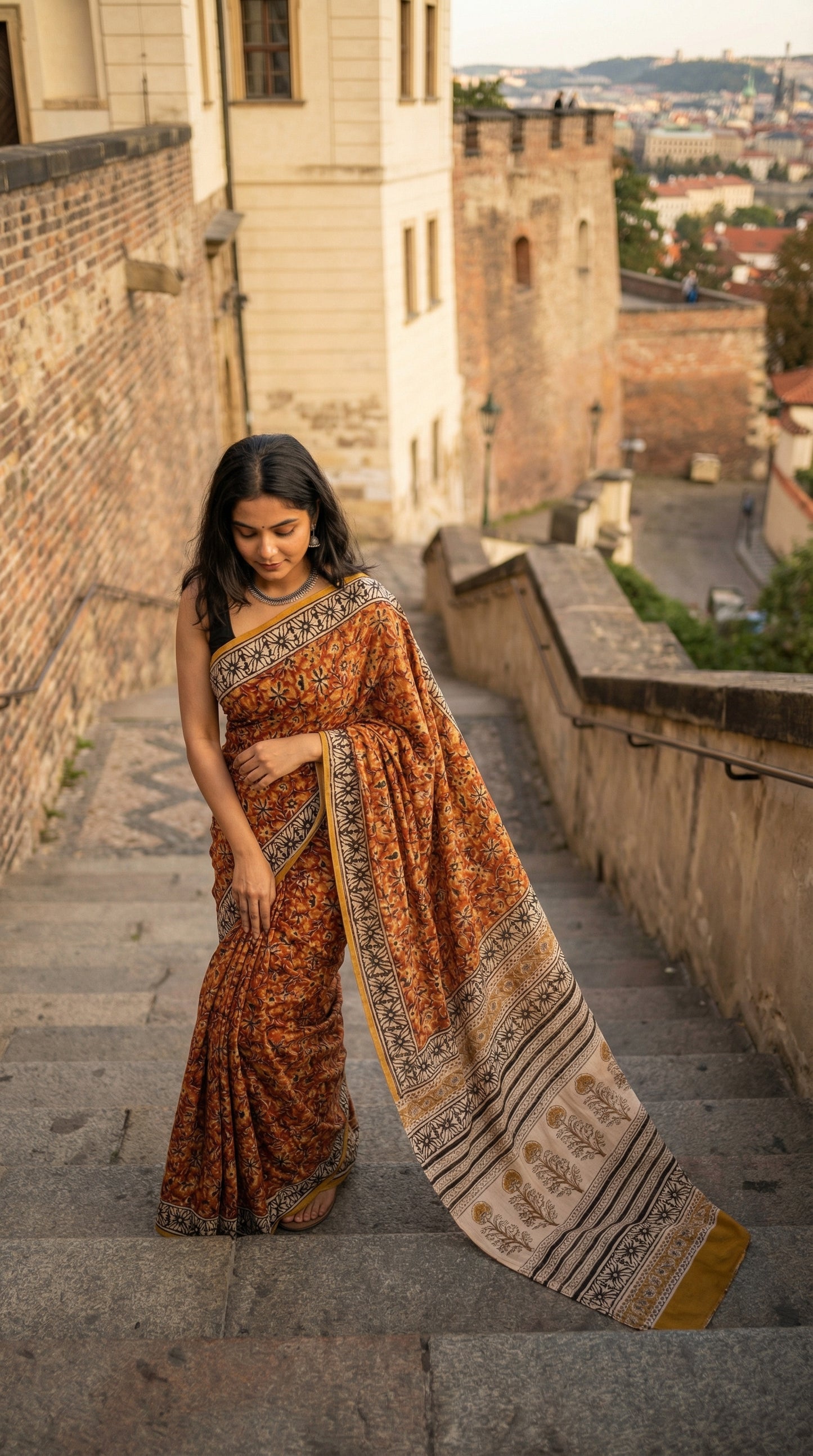 Woman wearing a Ajrakh Modal Silk Saree – Reddish-Orange with Black & White Border and Golden Pallu Motifs with intricate patterns standing on stone steps with a castle-like building in the background.