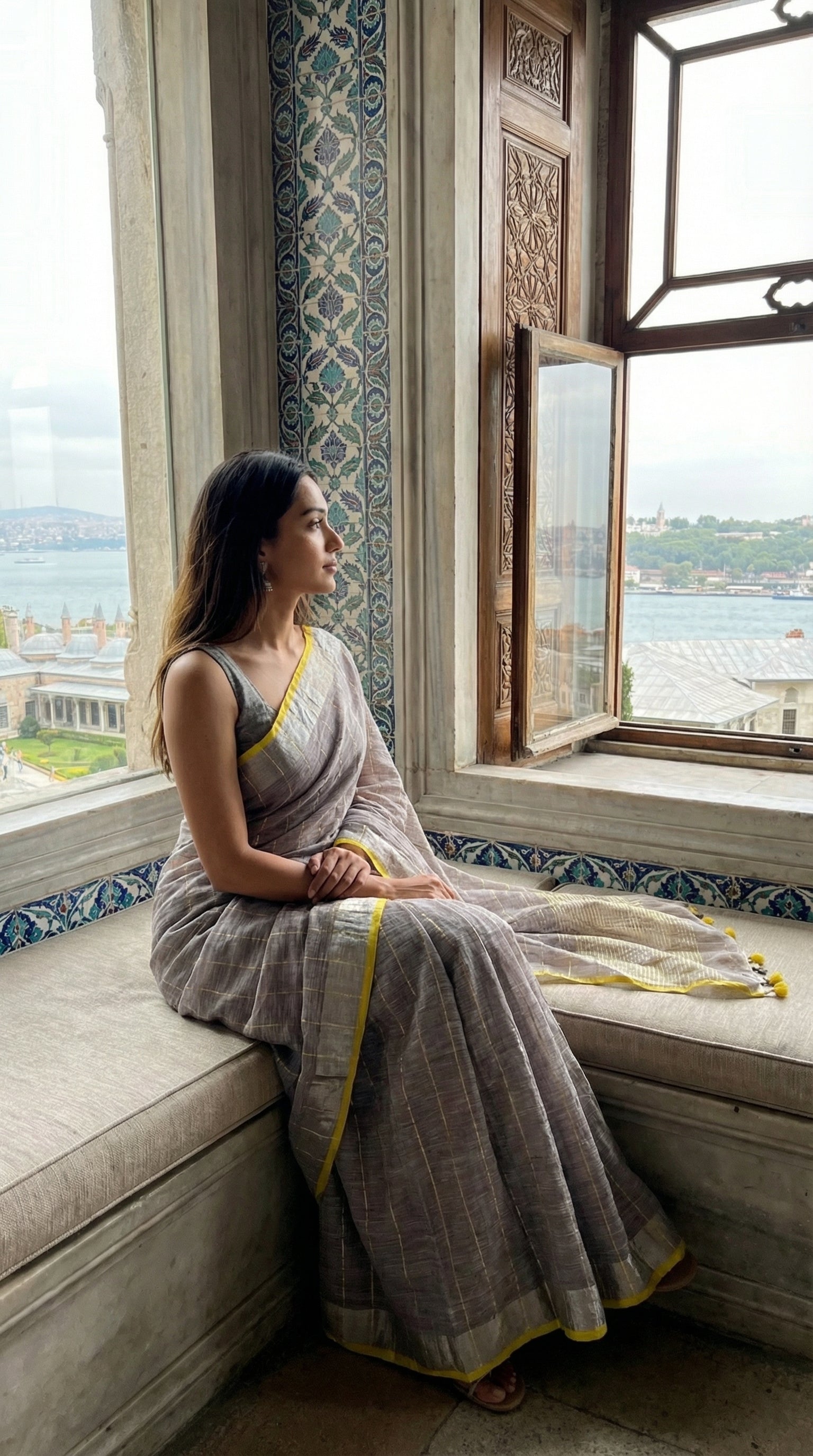 Woman in a ash-grey-cotton-linen-saree, sitting on a window sill with a scenic view.