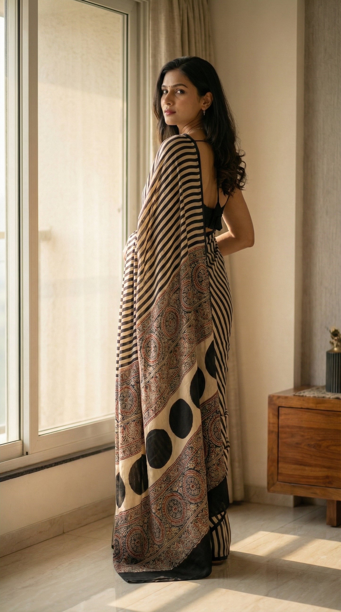 Woman in a beige and black modal silk striped saree with Ajrakh, standing by a window in a room with wooden furniture.