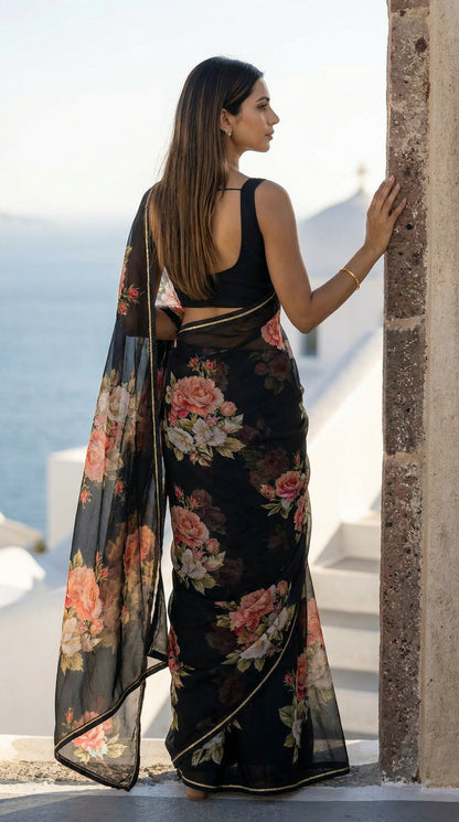 Woman wearing a black chiffon saree with floral prints, standing by a stone wall with ocean view.