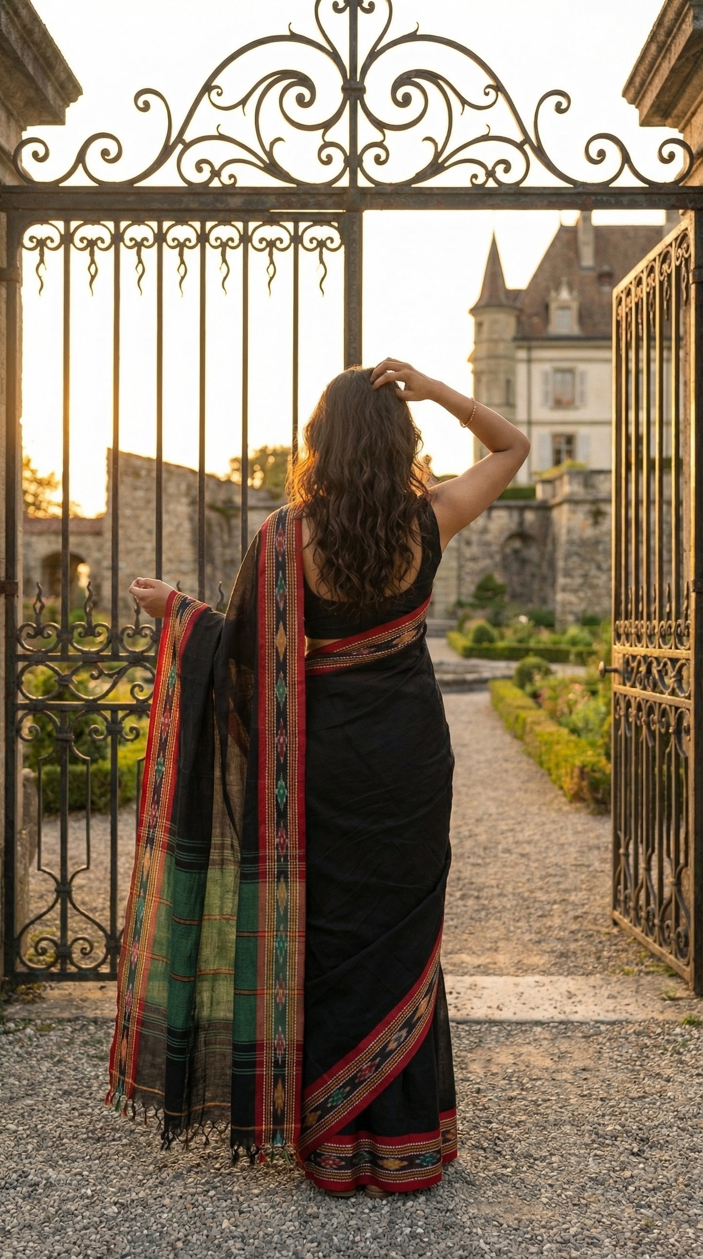 Woman in a black-mul-cotton-ikat-saree, standing in front of an ornate iron gate with a castle in the background.