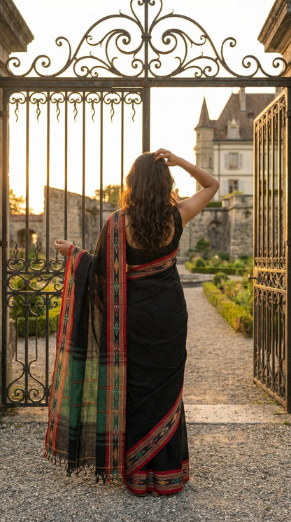 Woman in a black-mul-cotton-ikat-saree, standing in front of an ornate iron gate with a castle in the background.