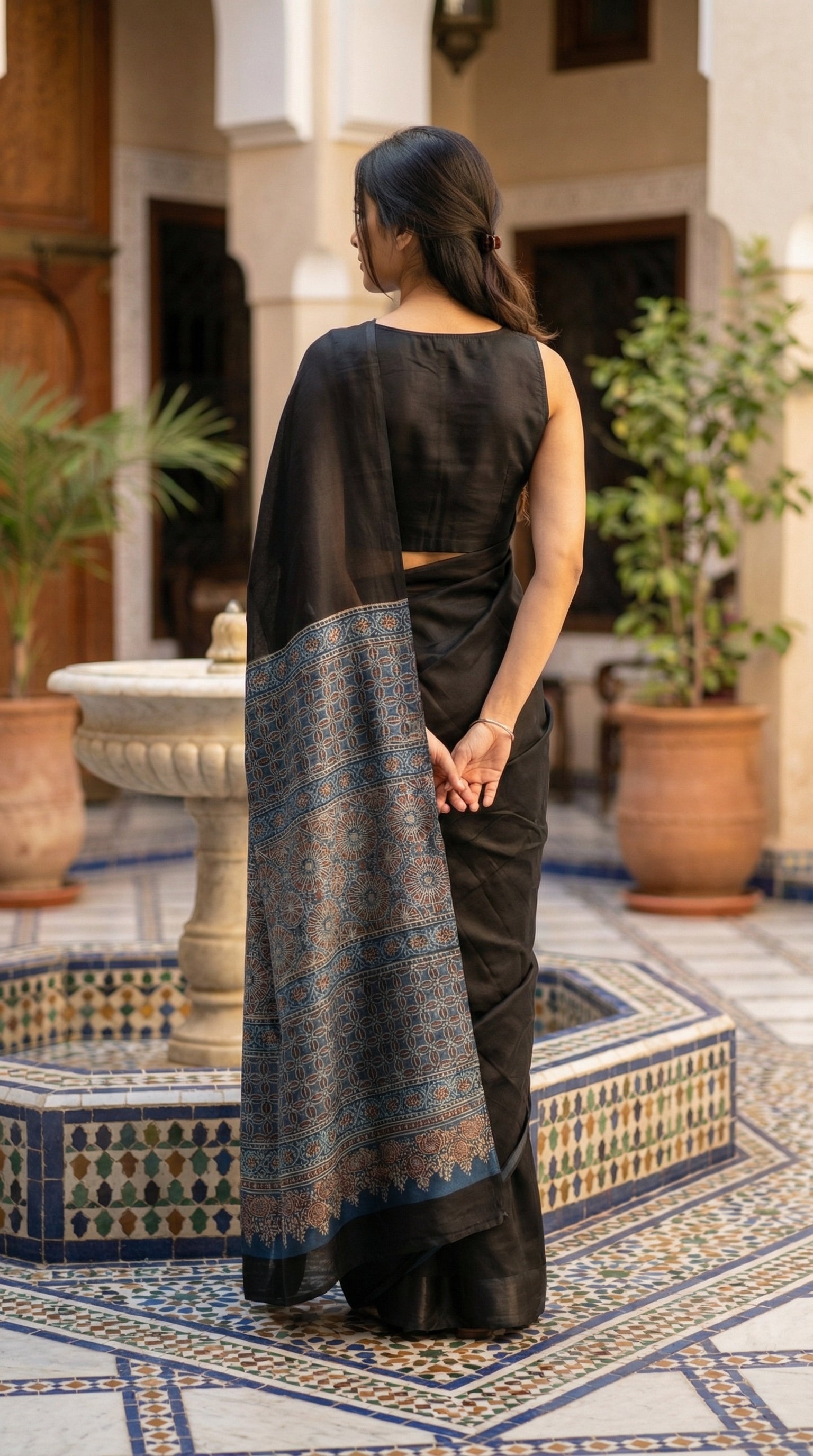 Woman in a black modal silk saree with intricate blue Ajrakh border detailing, standing in a decorative courtyard.