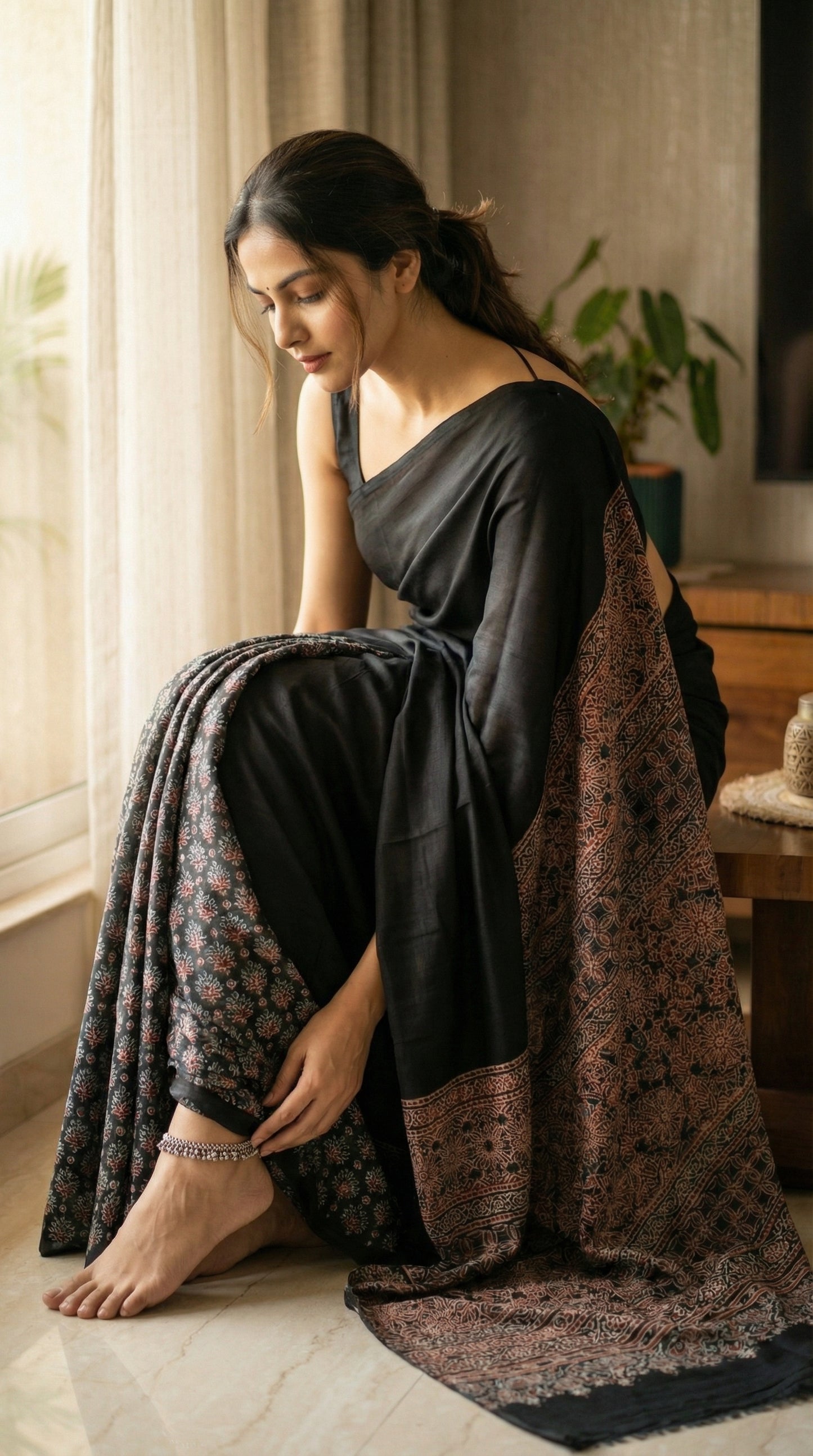 Woman in a black pure modal silk saree with intricate Ajrakh printed pallu, adjusting her anklet in her living room.