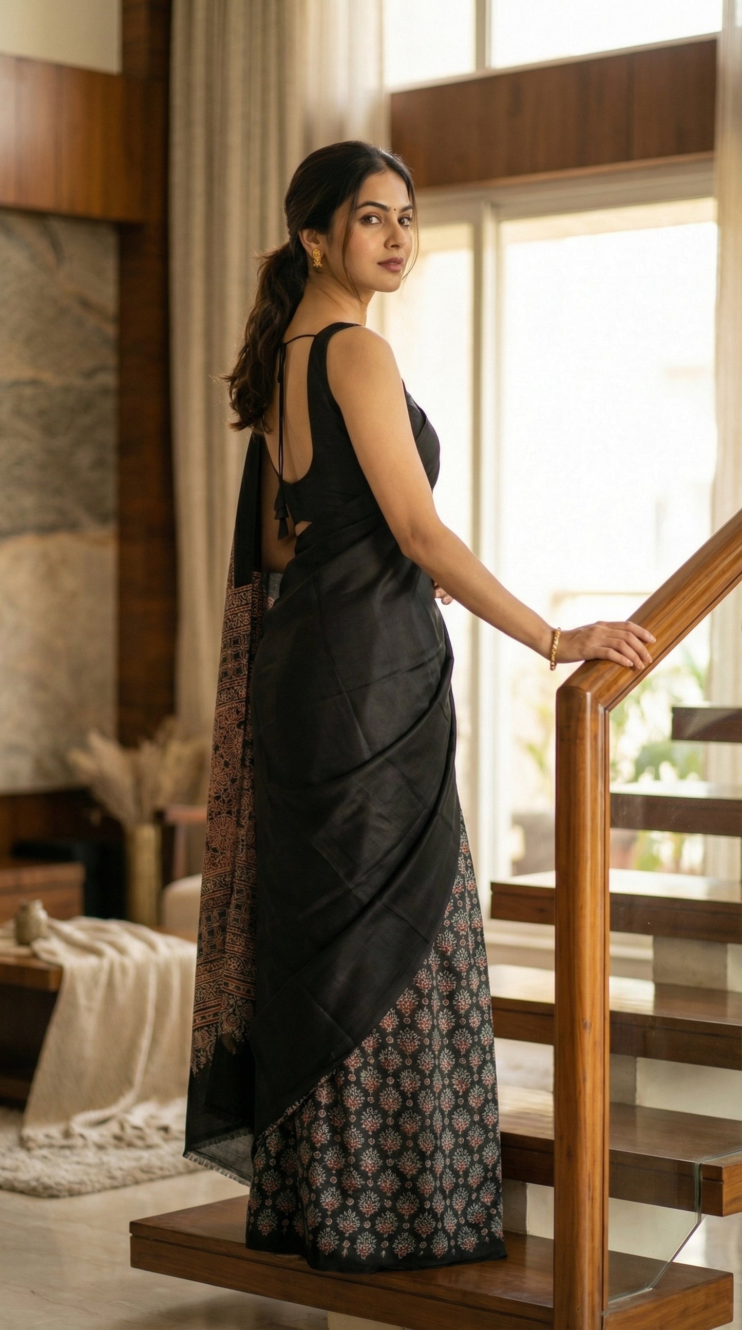Woman in a black pure modal silk saree with intricate Ajrakh printed pallu, standing on a staircase in a well-lit room.