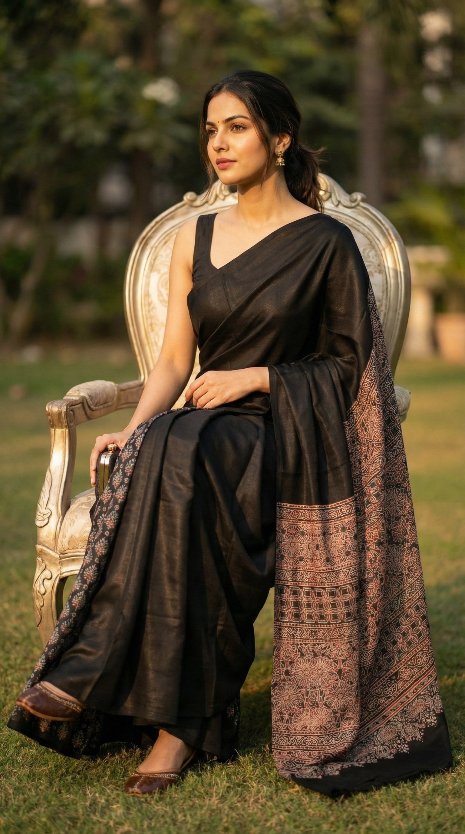 Woman in a black pure modal silk saree with intricate Ajrakh printed pallu, sitting outdoors on a chair.