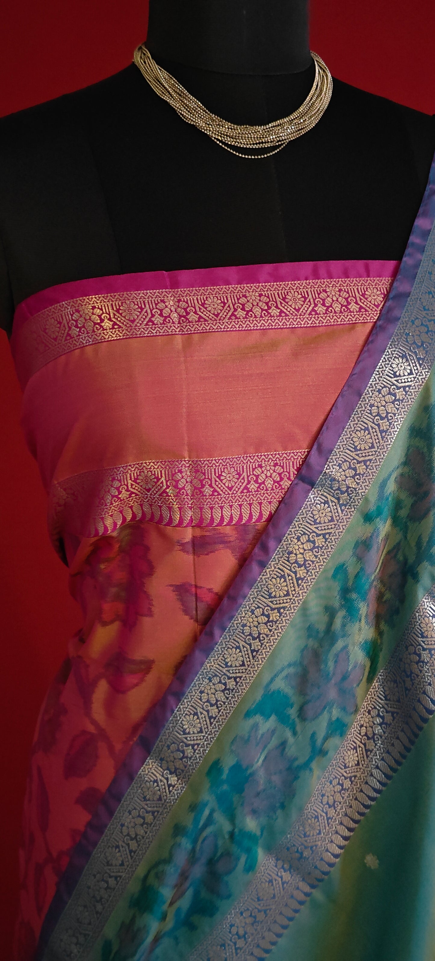 Blue blended silk saree with broad golden zari border and smooth finish on a mannequin with a vase of flowers against a red background.