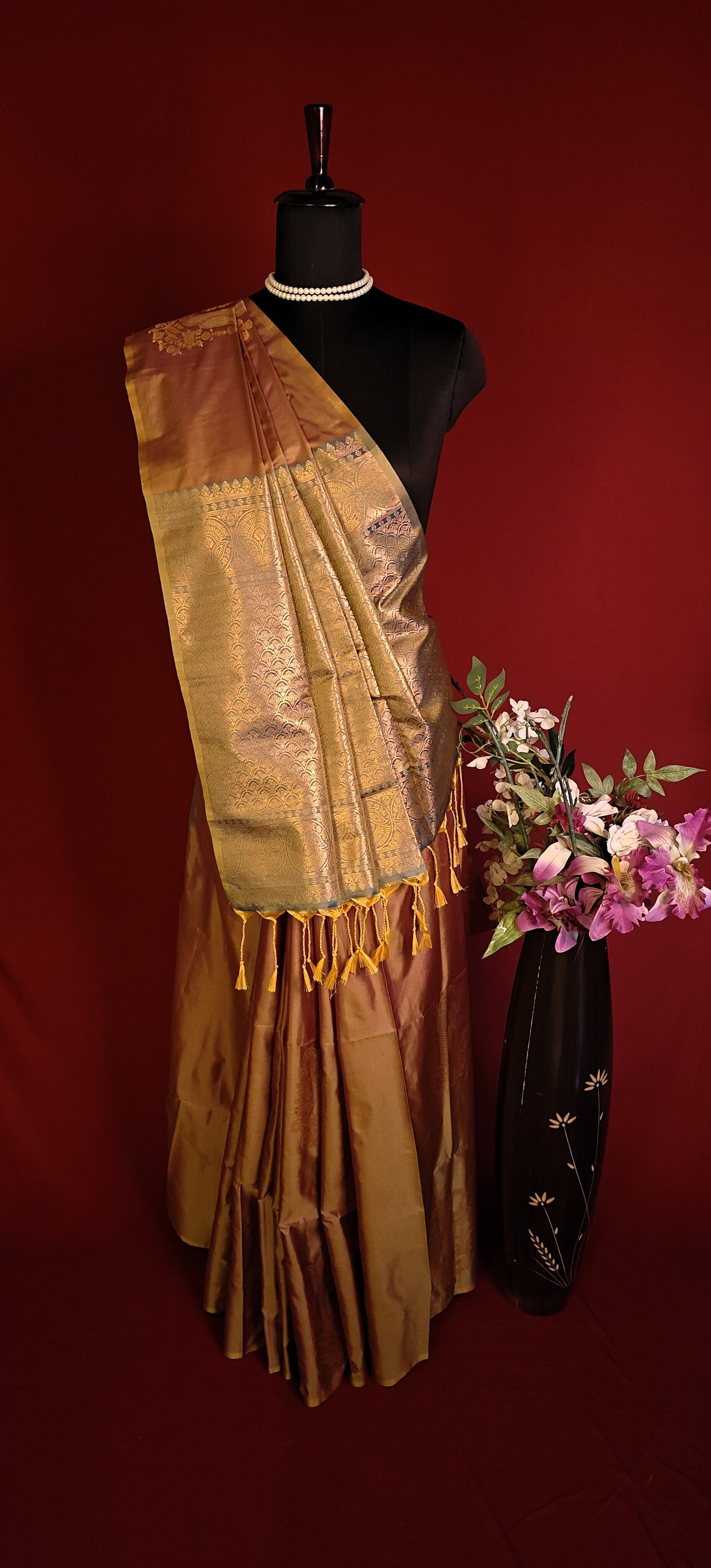 Copper brown blended silk saree with golden zari border and scattered butis, styled on mannequin with a vase of flowers on a red background.