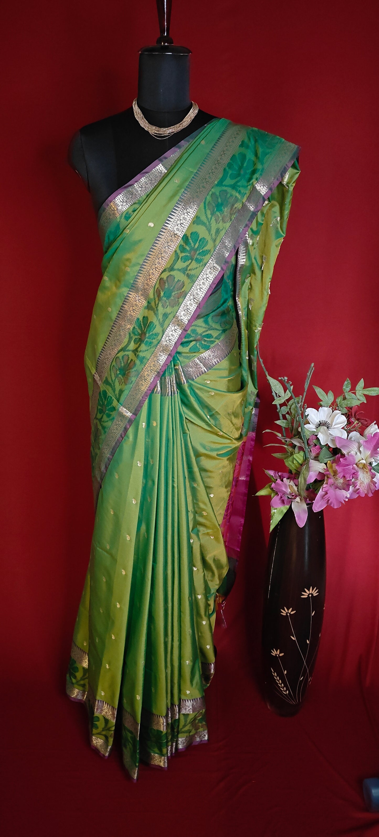 Green blended silk saree with elegant zari border and festive drape on a mannequin along with a flower vase against red background.