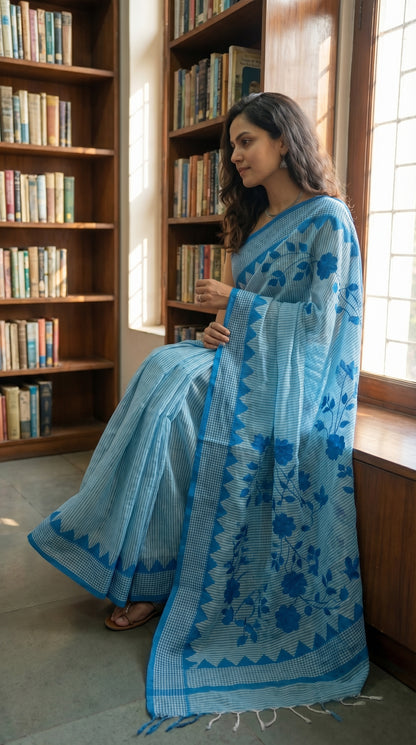 Woman in a blue-floral-cotton-applique-saree, standing in a room with bookshelves and a window.