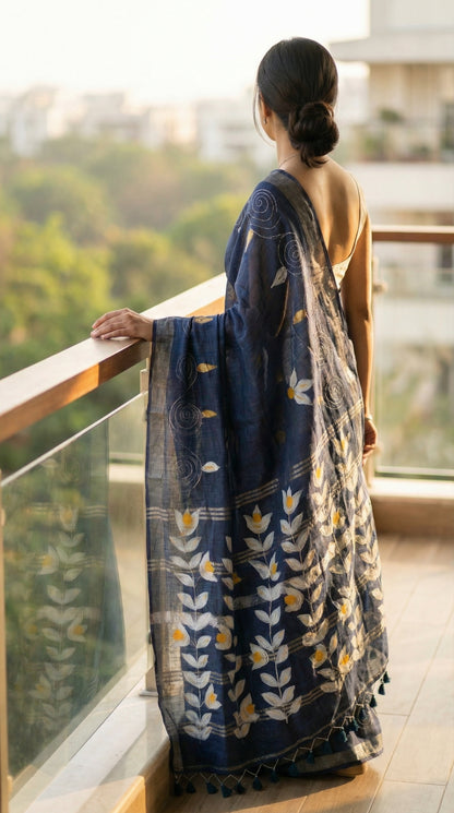 Woman in a charcoal grey hand painted cotton linen saree with floral motifs and tassel pallu, standing on a balcony overlooking greenery.