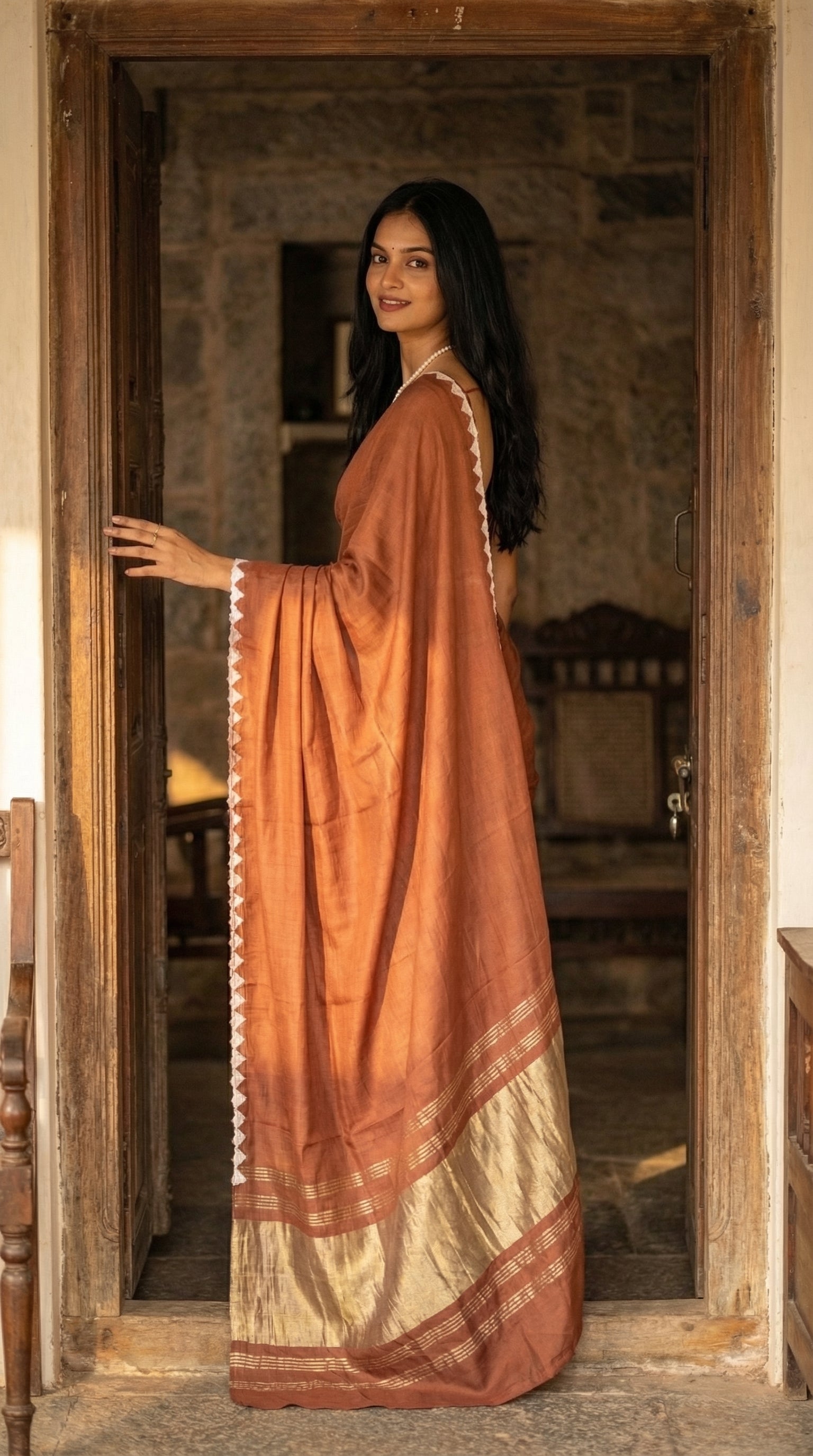 Woman wearing a chocolate-brown-modal-silk-saree-lagdi-patta-pallu, standing at the entrance of an old home.