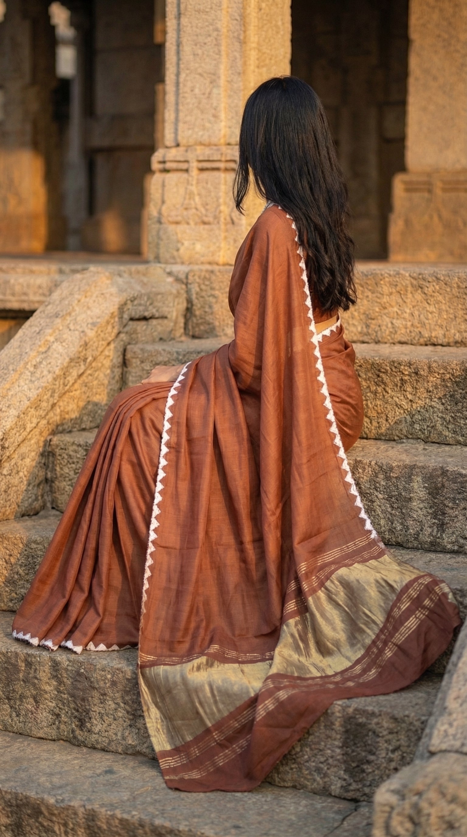 Woman wearing a chocolate-brown-modal-silk-saree-lagdi-patta-pallu, sitting on concrete chairs in an architectural setup.